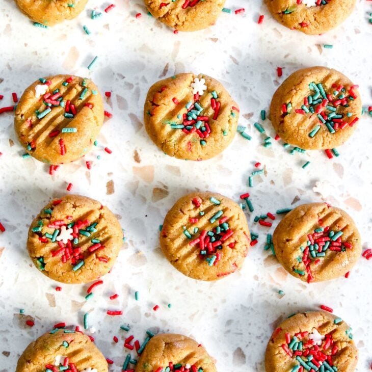 This is vertical image of cookies on a white terrazzo surface. The cookies are lined up in a 3x4 rows and have red, green and snowflake sprinkles on top. The tops of the cookies have lines on them indicating they have been pressed down with the back of a fork. More sprinkles are on the surface around the cookies.