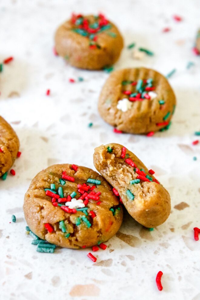 This is vertical image focusing on two cookies on a white terrazzo surface. One cookie has a bite taken out of it and is leaning against another cookie. More cookies are blurred in the background. The tops of the cookies have lines on them indicating they have been pressed down with the back of a fork. More sprinkles are on the surface around the cookies.