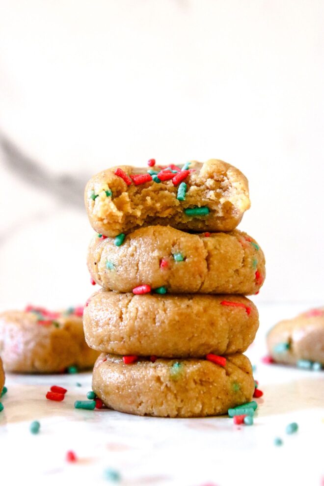 This is a stack of four no bake cookies. The cookies sit on a white surface with a white background. A bite is taken out of the top cookie and all the cookies have red and green sprinkles in and on top of them. More cookies are blurred around the stack of cookies.
