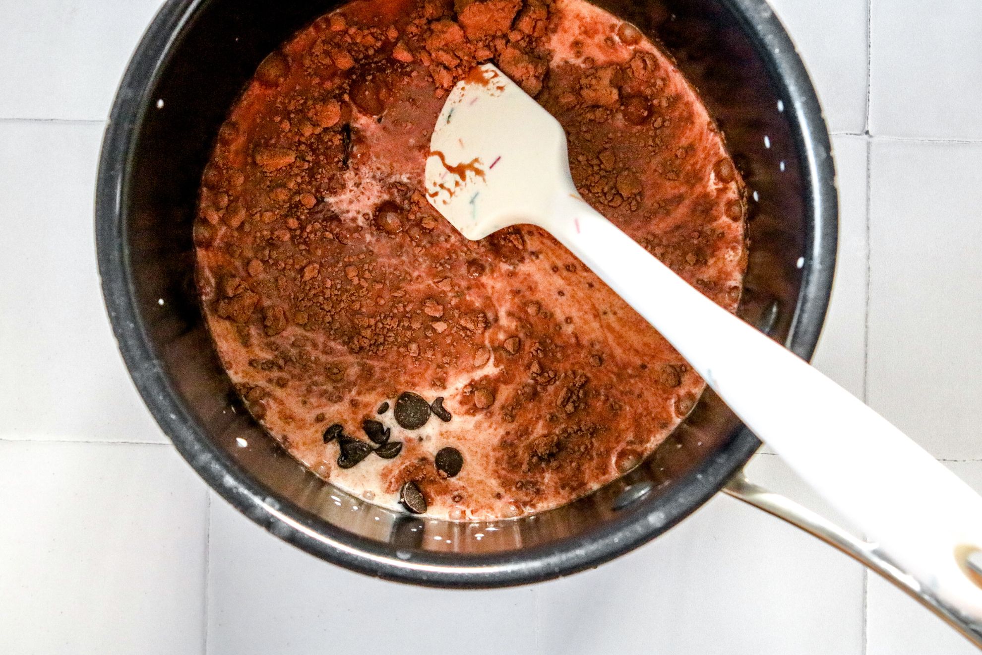 This is an overhead horizontal image of a black and silver pot with milk, cocoa powder, and chocolate chips in it. The pot sits on a white square tile surface. A white spatula is in the pot with the handle leaning against the side of the pot and pointing to the bottom right corner of the image.