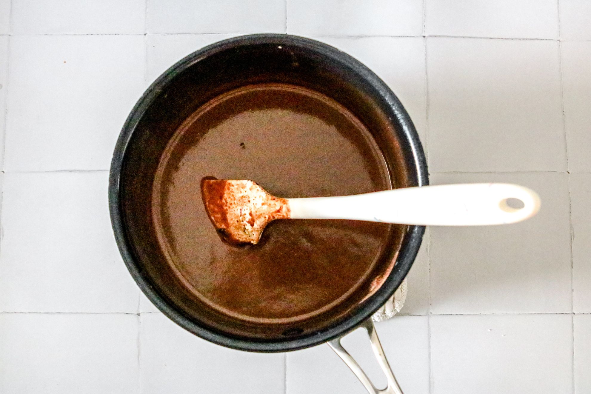 This is an overhead horizontal image of a black and silver pot with a chocolate mixture in it. The pot sits on a white square tile surface. A white spatula is in the pot with the handle leaning against the side of the pot and pointing to the middle right side of the image.