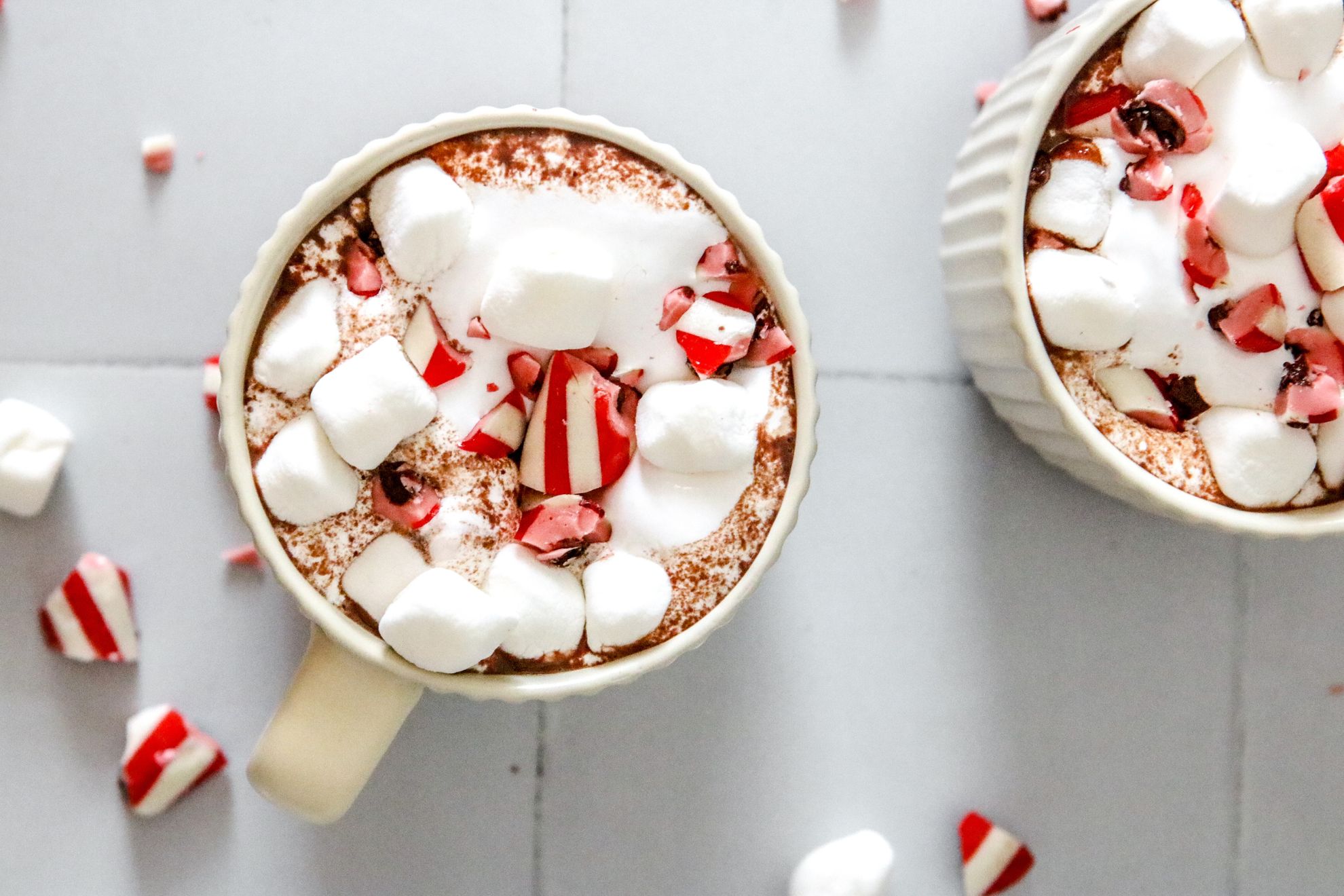 This is an overhead horizontal image of two mugs sitting on a white square tile surface. The image focuses on one mug and another mug is in the top right corner of the image. In the mug is fluff, mini marshmallows and crushed candy canes. More crushed candy canes and mini marshmallows are scattered around the mugs.