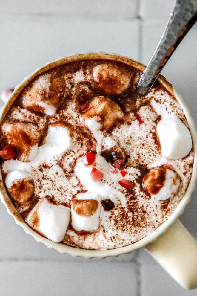 This is an overhead vertical image of a mug sitting on a white square tile surface. In the mug is fluff, mini marshmallows, chocolate and crushed candy canes. More crushed candy canes and mini marshmallows are scattered around the mugs. A silver utensil is dipping into the mug with the handle pointing at the top right corner of the image.