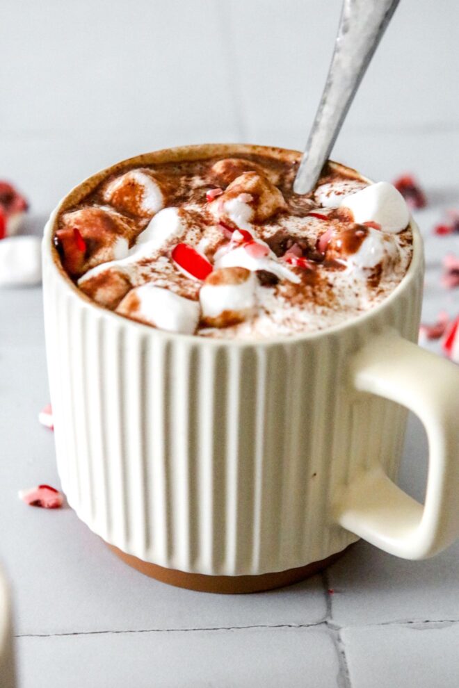 This is a vertical image looking at a mug from a side view. The mug is sitting on a white square tile surface. In the mug is fluff, mini marshmallows, chocolate and crushed candy canes. More crushed candy canes and mini marshmallows are scattered around the mugs. A silver utensil is dipping into the mug with the handle pointing at the top right corner of the image.