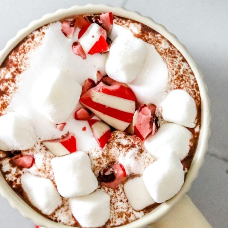 This is an overhead vertical image of a mug sitting on a white square tile surface. The image focuses on one mug and another mug is in the top right corner of the image. In the mug is fluff, mini marshmallows, hot chocolate, and crushed candy canes. More crushed candy canes and mini marshmallows are scattered around the mugs.