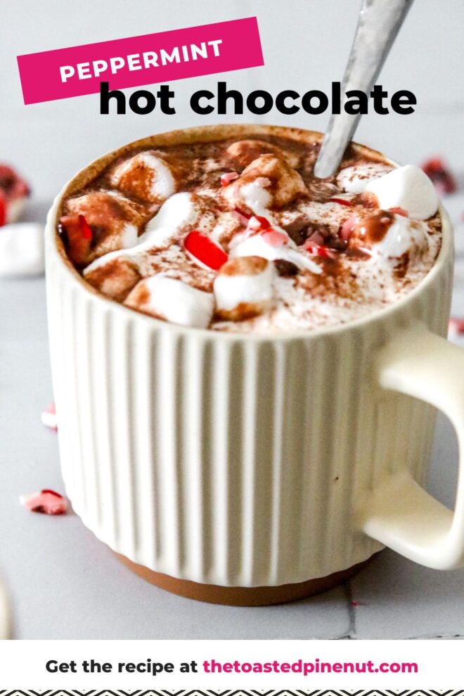This is a vertical image looking at a mug from a side view. The mug is sitting on a white square tile surface. In the mug is fluff, mini marshmallows, chocolate and crushed candy canes. More crushed candy canes and mini marshmallows are scattered around the mugs. A silver utensil is dipping into the mug with the handle pointing at the top right corner of the image. Text overlay reads "peppermint hot chocolate" at the top and "get the recipe at thetoastedpinenut.com"