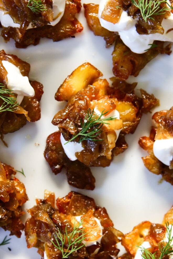 This is a vertical overhead image focusing on a crispy roasted smashed potato on a white plate. The center potato is topped with sour cream, caramelized onions, and a small bit of fresh dill. More crispy potatoes with the same toppings are on the plate around the center potato in focus.