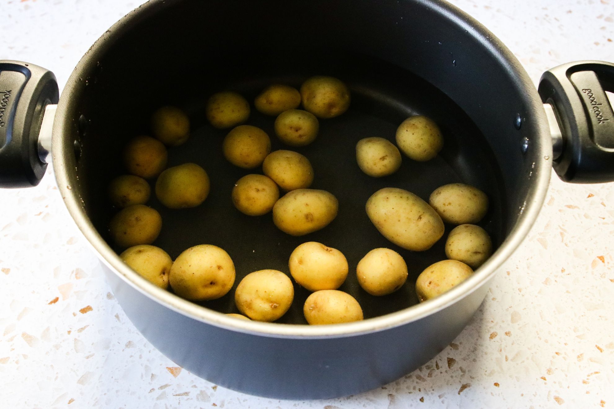 This is a horizontal image looking at a large pot with baby potatoes and water from an angled, overhead view. The pot sits on a white terrazzo surface.