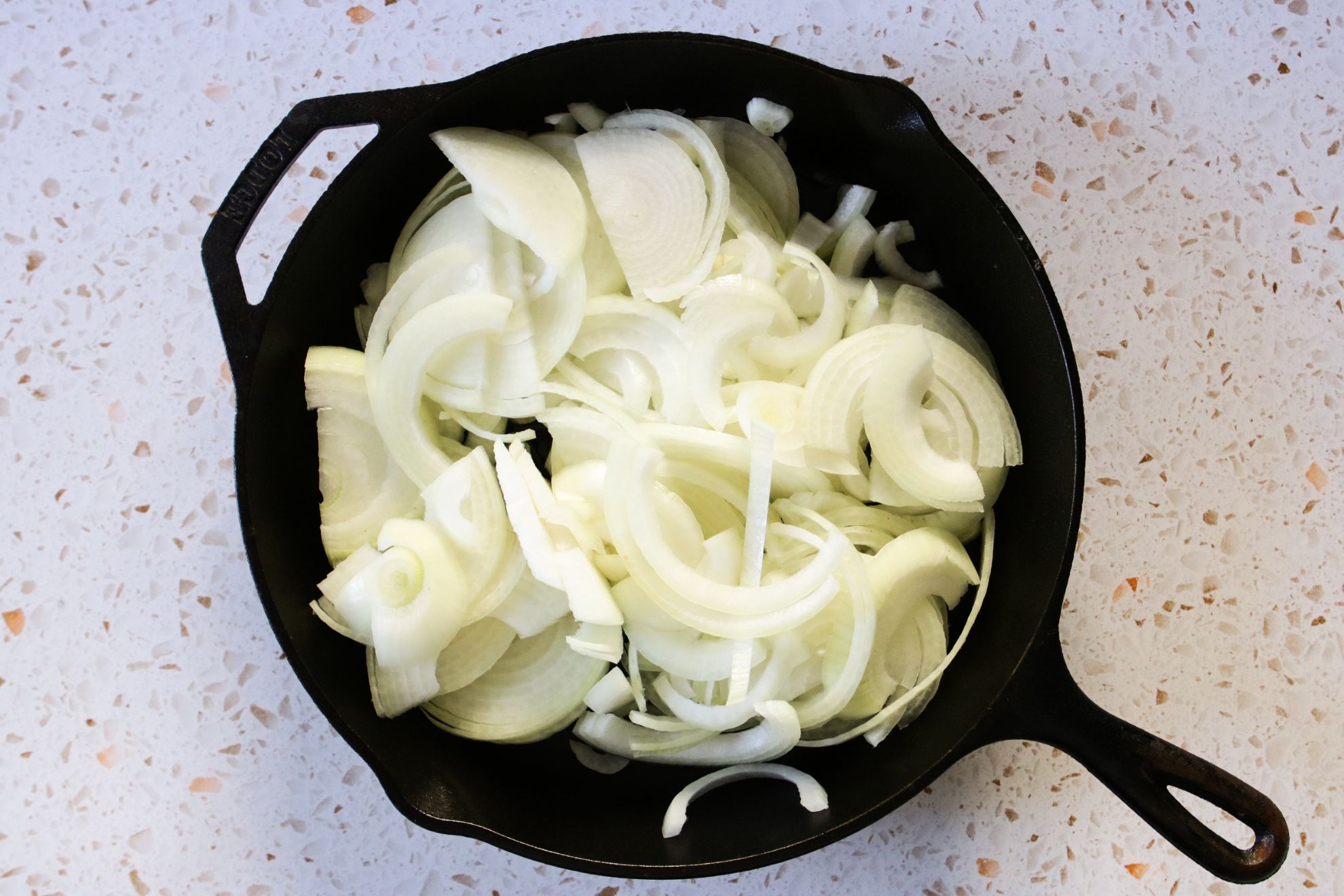 This is an overhead horizontal image of a cast iron skillet with raw sliced onions. The skillet sits on a whiter terrazzo surface.