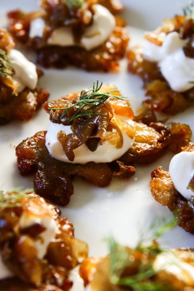 This is a vertical image focusing on a crispy roasted smashed potatoes on a white plate. The vantage point is from the side. The center potato is topped with sour cream, caramelized onions, and a small bit of fresh dill. More crispy potatoes with the same toppings are on the plate blurred around the center potato in focus.