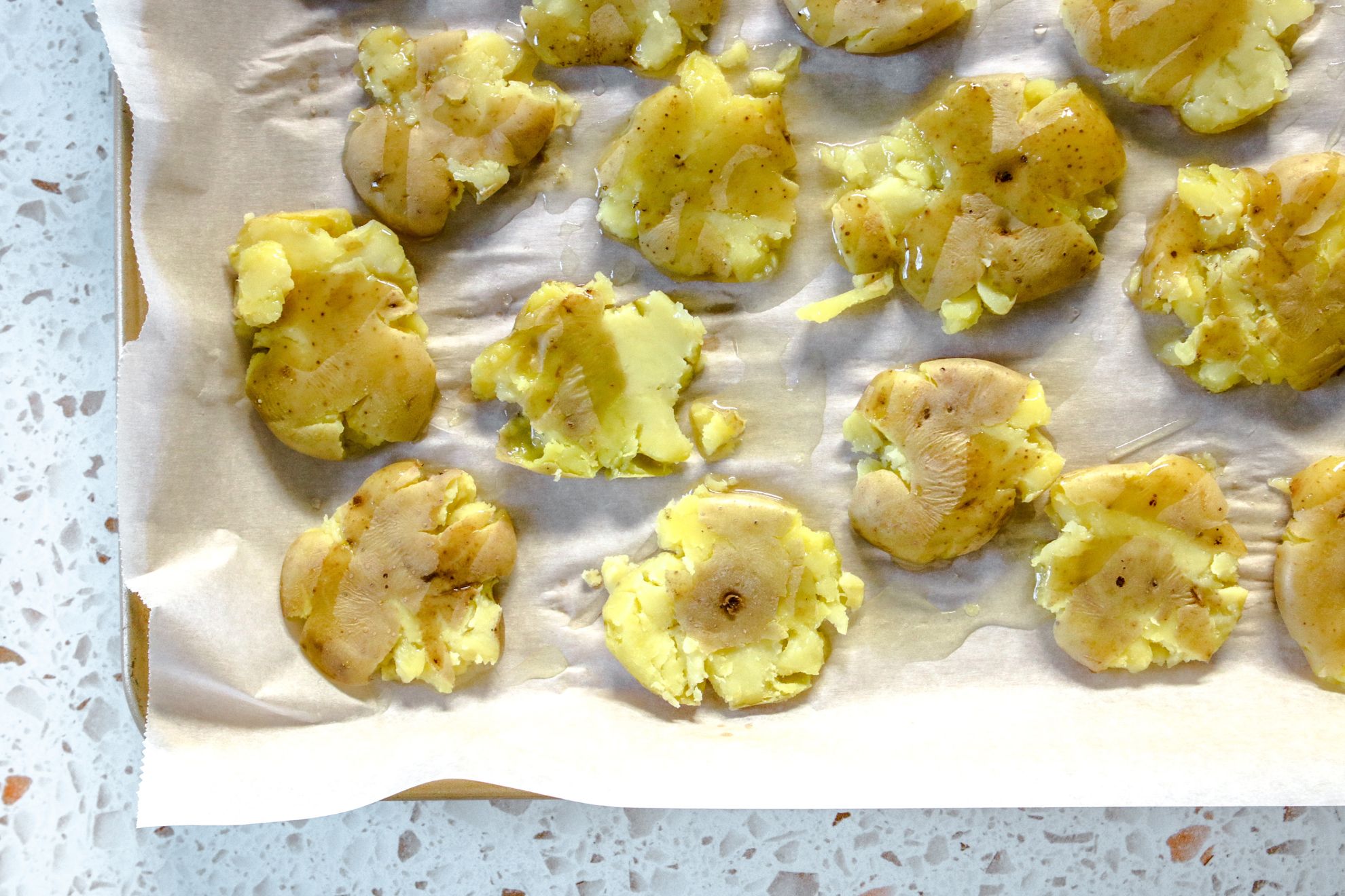 This is an overhead horizontal view of a gold rimmed baking sheet lined with parchment paper. On the baking sheet are smashed potatoes drizzled with oil. The baking sheet sits on a white terrazzo surface.
