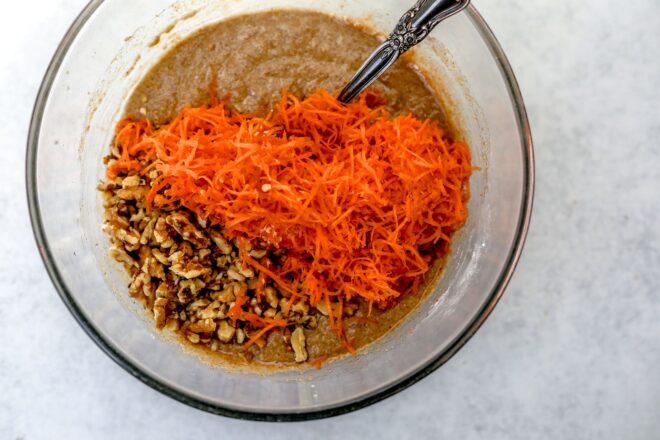 This is an overhead horizontal image of a glass bowl with mixed cake batter and topped with shredded carrots and chopped walnuts. The bowl sits on a light grey surface. A silver spoon is in the batter and leaning against the side of the bowl.