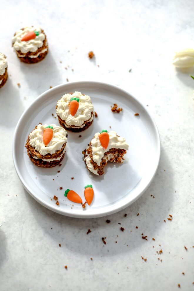 This is a vertical image looking at a white plate from an overhead angle. On the plate are three mini carrot cakes. One mini carrot cake has a bite taken out of it. Candy carrots are on the plate next to the cakes and each cake has a candy carrot on it as well. The plate sits on a light grey surface with more mini cakes to the top left corner of the image and white tulips are at the top right corner of the image.