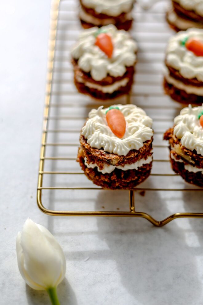 This is a vertical image of mini layered carrot cakes on a cooling rack. The cooling rack sits on a white surface. The cakes have a layer of cream cheese frosting in the middle and frosting on the top with a candy carrot. The image focuses on on cake but more are blurred around the cake on the cooling rack. A single white tulip is in the bottom left corner of the image.
