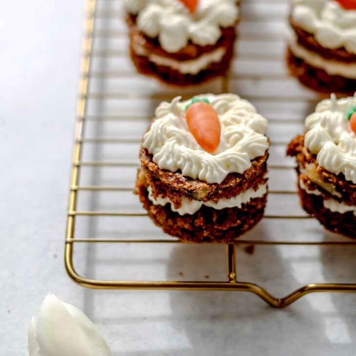 This is a vertical image of mini layered carrot cakes on a cooling rack. The cooling rack sits on a white surface. The cakes have a layer of cream cheese frosting in the middle and frosting on the top with a candy carrot. The image focuses on on cake but more are blurred around the cake on the cooling rack. A single white tulip is in the bottom left corner of the image.