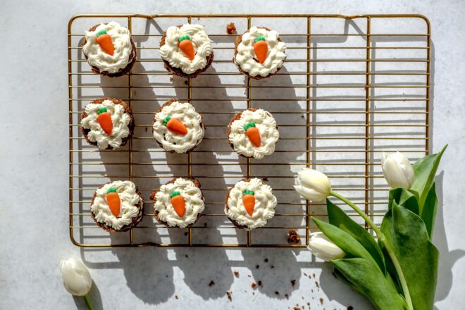 This is an overhead horizontal image of a gold cooling rack on a light grey surface. On the cooling rack are nine mini carrot cakes topped with cream cheese frosting an carrot candies. Three white tulips are in the bottom right corner and one tulip is to the bottom left.