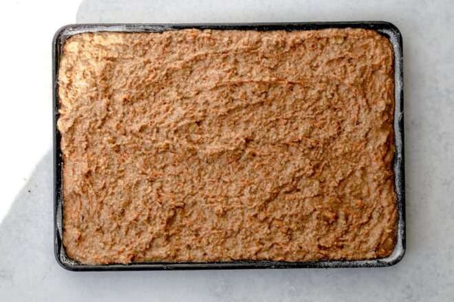 This is an overhead horizontal image of a sheet pan with carrot cake batter spread out in it. The sheet pan sits on a light grey surface.