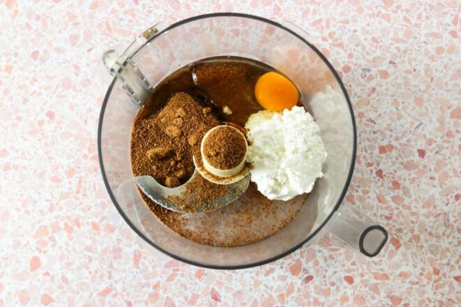 This is an overhead horizontal image of a food processor with cottage cheese, coconut sugar, and an egg in it. The food processor sits on a pink and white terrazzo surface.