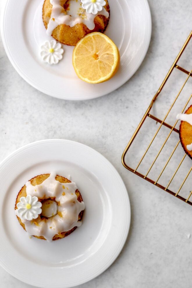 This is an overhead vertical image of two small white plates with mini bundt cakes on them. The mini bundt cakes have a white icing glaze and sugar flower decorations. The plate to the top of the image has half of a lemon on it. A gold cooling rack with part of a a mini bundt cake is coming from the right center of the image.
