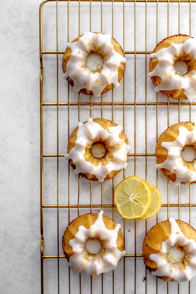 This is an overhead vertical image looking at a cooling rack with mini bundt cakes on it. The gold cooling rack sits on a white surface. All the mini bundt cakes are topped with a white glaze. Two slices of lemon are on the cooling rack in between the mini bundt cakes.