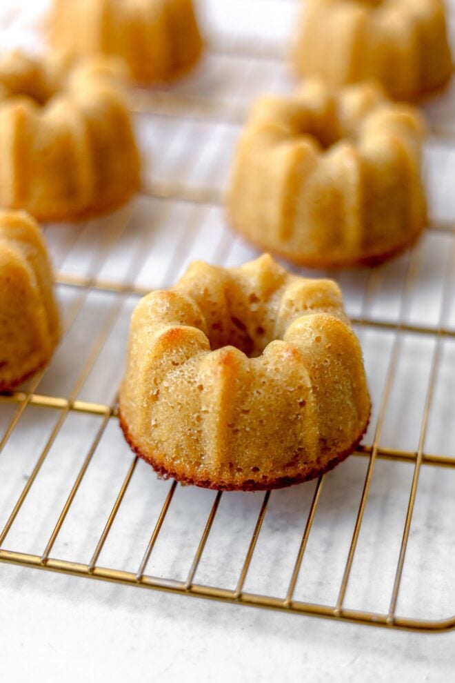 This is a vertical image looking at a cooling rack with mini bundt cakes on it from the side. The gold cooling rack sits on a white surface.