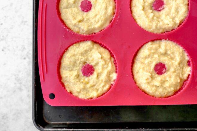 This is an overhead horizontal image of a pink silicone mini bundt pan on a rimmed baking sheet. The rimmed pan is on a light grey surface. In the silicon pan is raw batter.