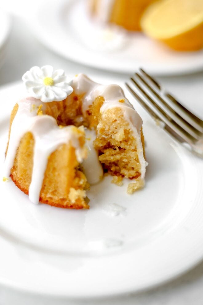 This is a vertical image of a mini bundt cake on a small white plate. The image is looking at the cake from the side. The cake has a white glaze on top and dripping down the sides and middle. A white sugar flower is on top of the bundt cake and a fork is on the right side of the image leaning against the side of the plate. Another plate with a mini bundt cake and a lemon wedge is on a small white plate blurred in the background.