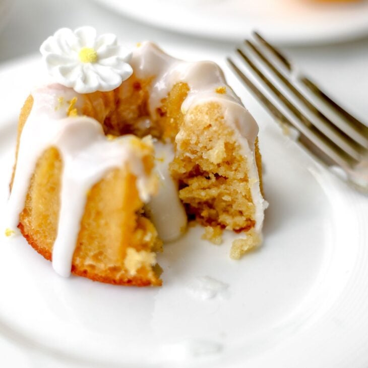 This is a vertical image of a mini bundt cake on a small white plate. The image is looking at the cake from the side. The cake has a white glaze on top and dripping down the sides and middle. A white sugar flower is on top of the bundt cake and a fork is on the right side of the image leaning against the side of the plate. Another plate with a mini bundt cake and a lemon wedge is on a small white plate blurred in the background.