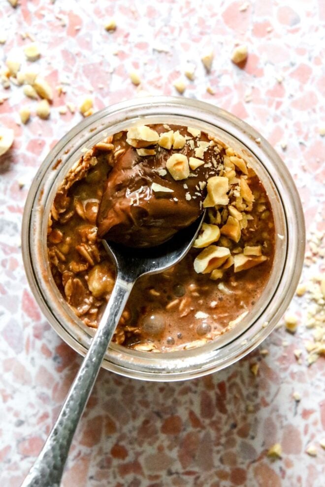 This is an overhead vertical image of a glass jar with chocolate overnight oats in it. A silver spoon is dipping into the oats with the handle leaning against the bottom left side of the jar. Crushed nuts are on top of the chocolate oats and on the pink terrazzo surface around the jar.