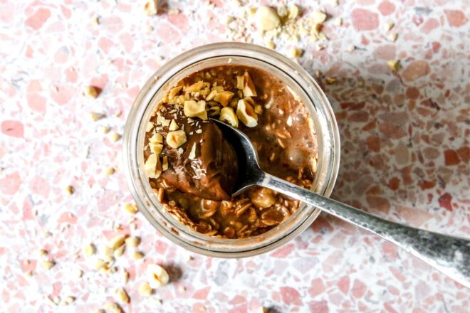 This is an overhead horizontal image of a glass jar with chocolate overnight oats in it. A silver spoon is dipping into the oats with the handle leaning against the right side of the jar. Crushed nuts are on top of the chocolate oats and on the pink terrazzo surface around the jar.