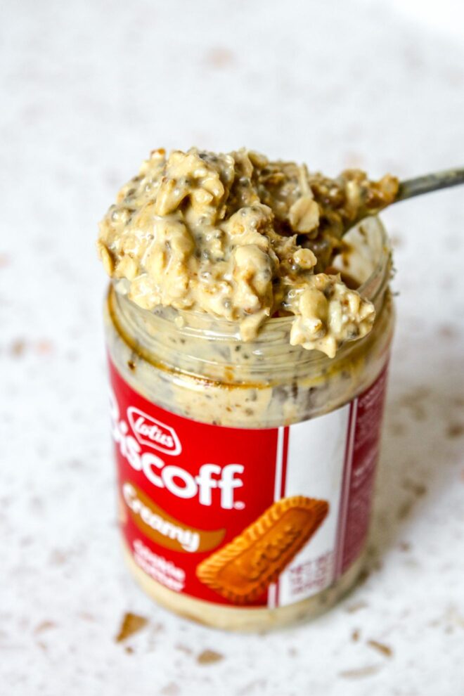 This is a vertical image looking at a labeled biscoff jar filled with overnight oats from an overhead, angled view. A spoon with a heaping spoonful of oats is leaning across the top of the jar. The jar sits on a white terrazzo surface with a white tile background.