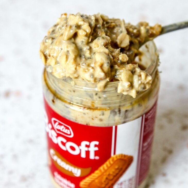 This is a vertical image looking at a labeled biscoff jar filled with overnight oats from an overhead, angled view. A spoon with a heaping spoonful of oats is leaning across the top of the jar. The jar sits on a white terrazzo surface with a white tile background.