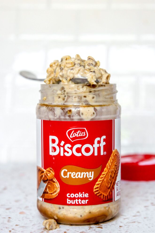This is a vertical image looking at a labeled biscoff jar filled with overnight oats from the side. A spoon with a heaping spoonful of oats is leaning across the top of the jar. The jar sits on a white terrazzo surface with a white tile background. A red plastic lid is on the surface behind the biscoff jar blurred in the background.