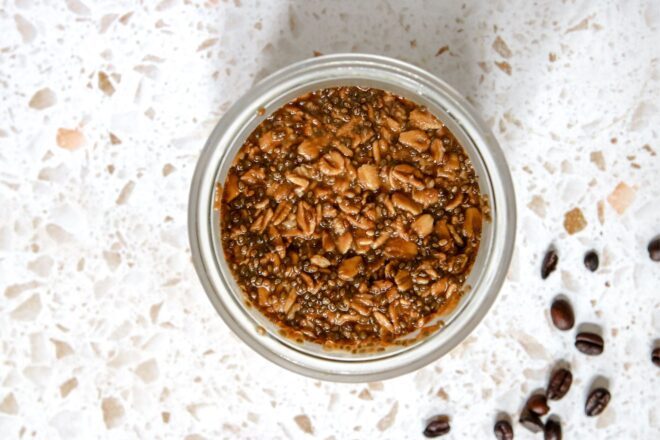 This is an overhead horizontal image of a glass jar with overnight oats in it. The jar sits on a white terrazzo surface with whole coffee beans to the bottom right corner of the image.