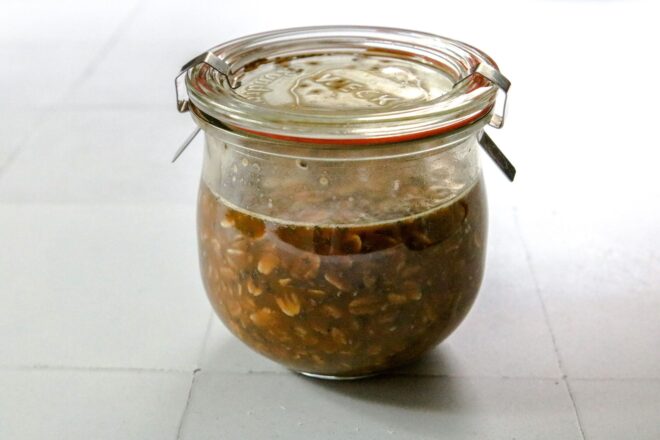 This is a horizontal image looking at a glass jar from the side. The jar sits on a white square tile surface. In the jar is a light brown liquid and oats. The jar has a glass lid secured with metal clasps.