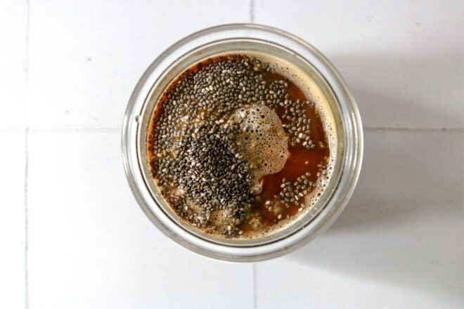 This is an overhead horizontal image of a glass jar with seeds, coffee, and sweetener in it. The glass jar sits on a white square tile surface.