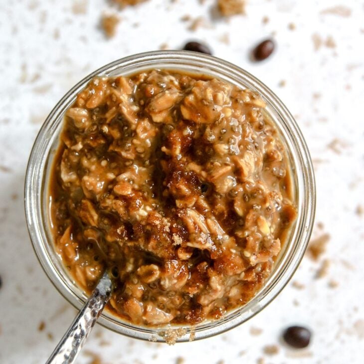 This is a vertical overhead image looking at a small glass jar. In the jar are deep brown colored overnight oats with a silver spoon sticking out and leaning against the left side of the jar, pointing to the bottom left corner of the image. The jar sits on a white terrazzo surface with a few whole coffee beans and brown sugar scattered around the jar.