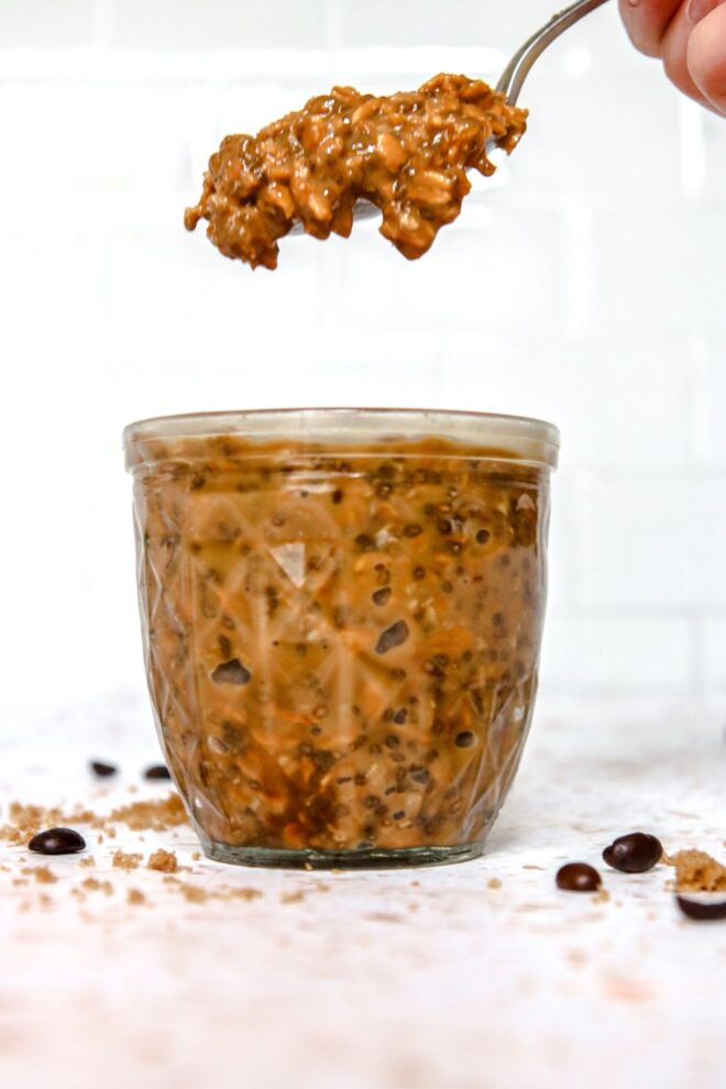 This is a vertical image looking at a small glass jar from the side. In the jar are deep brown colored overnight oats. The jar sits on a white terrazzo surface with a few whole coffee beans and brown sugar scattered around the jar. A hand holding a silver spoon with oats on it is coming in from the right top of the image directly above the glass jar.