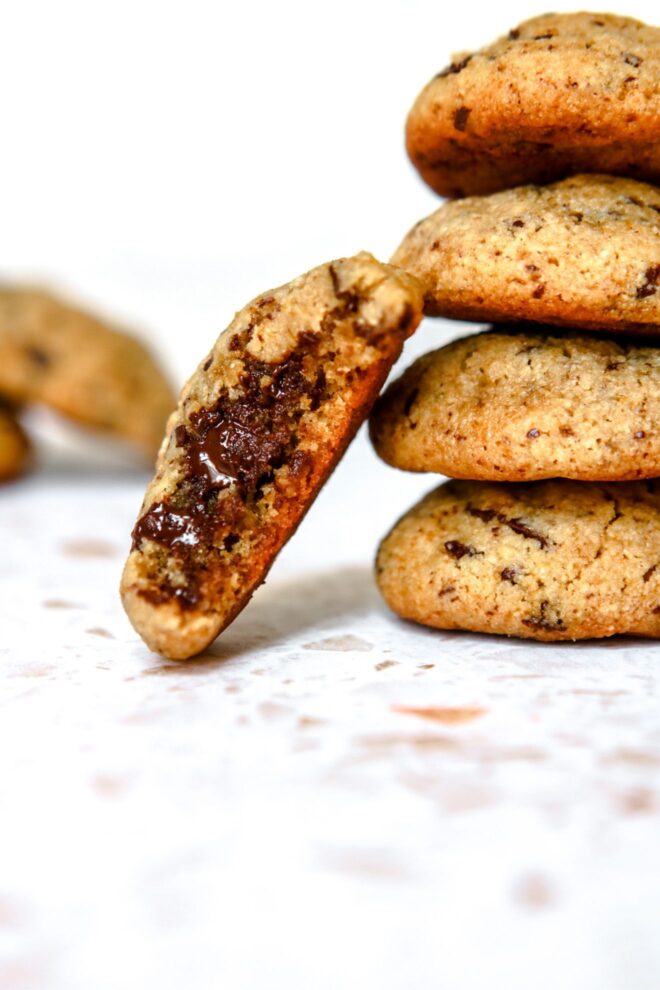 This is a vertical image looking at a stack of four chocolate chip cookies from the side. The stack sits on a white terrazzo surface. Another cookie has a bite taken out of it and is leaned up against the stack of four. More cookies are blurred in the background.