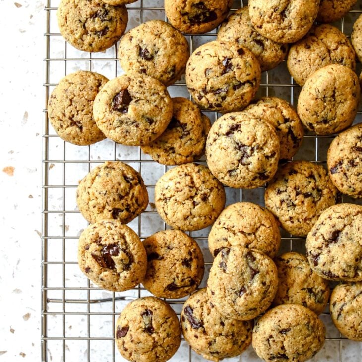 This is an overhead vertical image of a silver cooling rack with mini chocolate chip cookies piled on top. The image focuses on the bottom left corner of the cooling rack with the rest of the cooling rack out of frame. The cooling rack sits on a white terrazzo surface.
