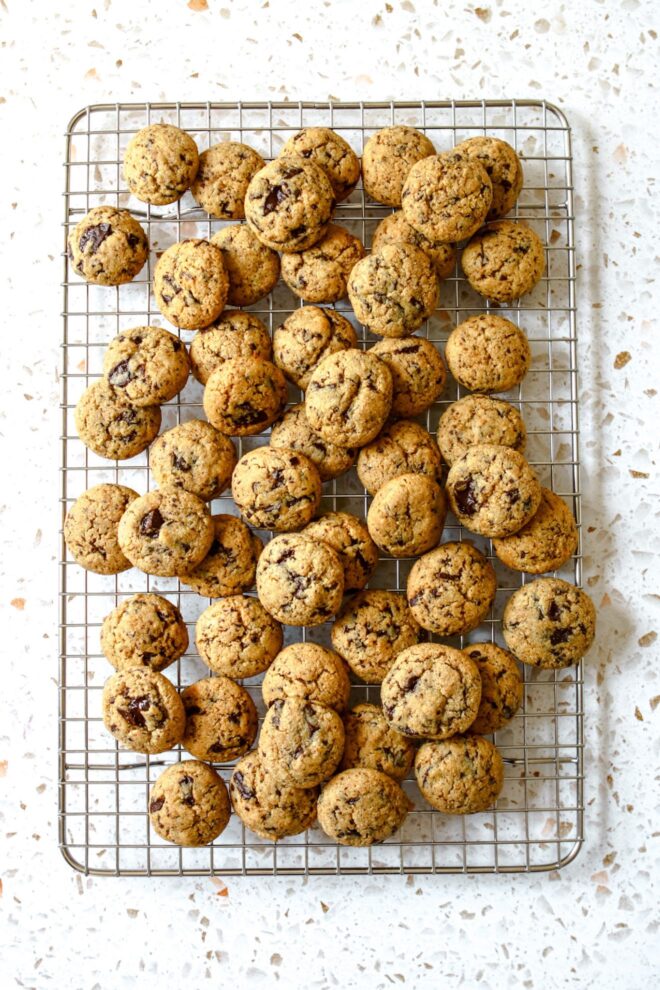 This is an overhead vertical image of a silver cooling rack with mini chocolate chip cookies piled on top. The cooling rack sits on a white terrazzo surface.