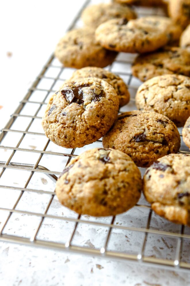 This is a vertical image looking at a silver cooling rack from the side. On the cooling rack are mini chocolate chip cookies. The image focuses on on cookie that has a melty chocolate chunk in the middle. The cookie leans against the other cookies. The cooling rack sits on a white terrazzo surface.