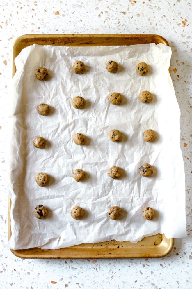 This is an overhead vertical image of a gold rimmed baking sheet with white parchment paper. On the parchment paper is a 4 x 5 grid of mini chocolate chip cookie dough balls. The baking sheet sits on a white terrazzo surface.