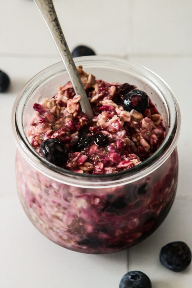 his is a vertical image looking at a glass jar with deep purple oats and blueberries in it from the side. A silver spoon is dipping into the oats with the handle leaning against the back rim of the jar pointing to the top left corner. The jar sits on a white tile surface with blueberries scattered around it.