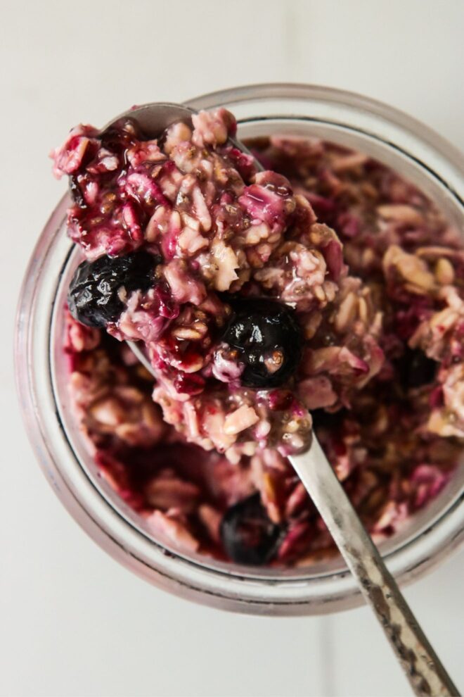 This is an overhead vertical image of a glass jar with deep purple oats and blueberries in it. A silver spoon has a heaping spoonful of oats in it and is leaning against the rim of the jar. The jar sits on a white tile surface.