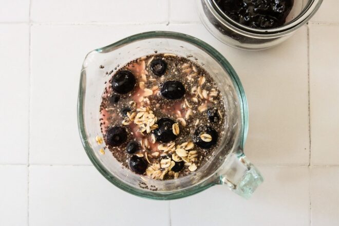 This is an overhead horizontal image looking into a liquid measuring cup with water, blueberries, oats and chia seeds in it. The measuring cup sits on a white square tile surface. A glass jar with blueberry jam is peeking in from the top right corner.