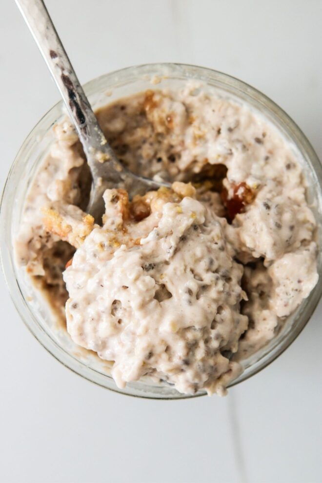 This is an overhead vertical image of a glass jar with a light pink oaty mixture with chia seeds. A silver spoon is in the jar scooping some of the mixture up and leaning against the side of the jar.