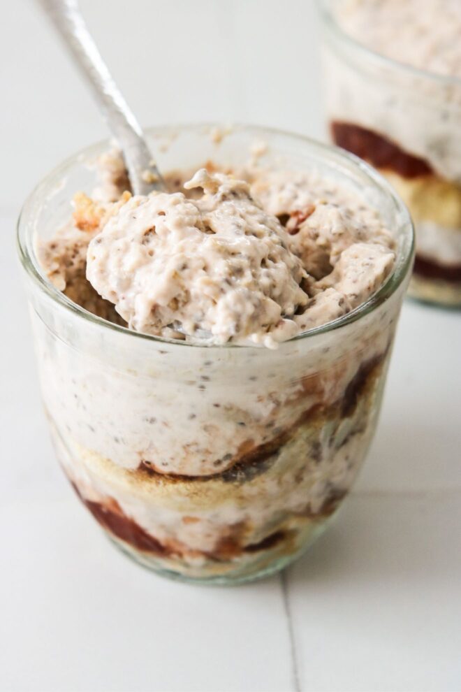 This is a vertical image looking at a glass jar from the side. In the glass jar are layers of graham cracker crumbs, strawberry jam, and a light pink almost white mixture with specks of seeds in it. A silver spoon is in the jar scooping up the oats and leaning against the side of the jar. Another glass jar with similar layers is behind the fro centered jar blurred in the background. The jars sit on a white square tile surface.