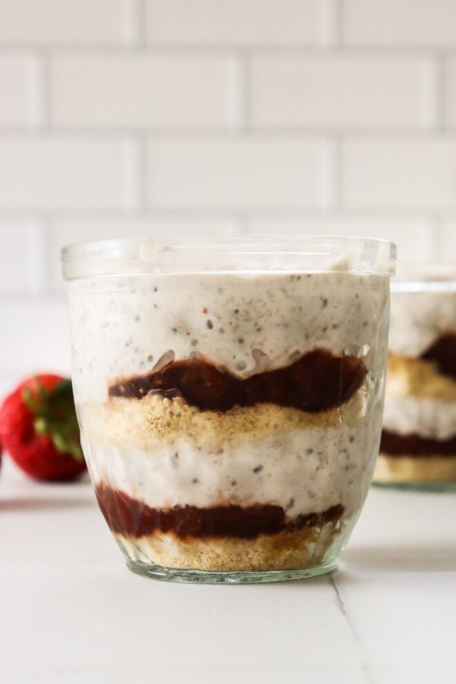 This is a vertical image looking at a glass jar from the side. In the glass jar are layers of graham cracker crumbs, strawberry jam, and a light pink almost white mixture with specks of seeds in it. Another glass jar with similar layers is behind the fro centered jar blurred in the background. The jars sit on a white square tile surface and a single strawberry is in the back left corner of the image.