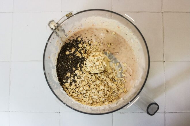 This is an overhead horizontal image of a food processor with a light pink mixture in it. On top of the light pink mixture are oats and chia seeds. The food processor sits on a white square tile surface.